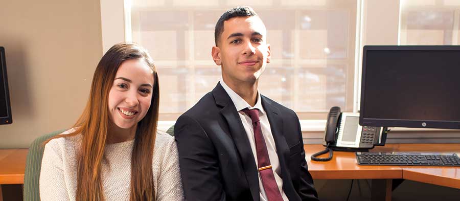 Caroline Fuentes '19 and Nolan Bebarta '20 sitting on buisness chairs with a computer and office phone on a desk in the background